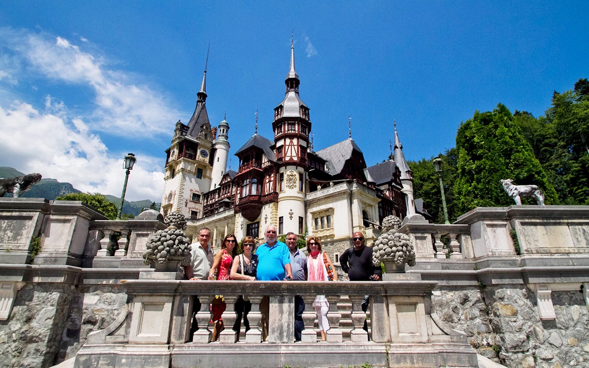 Guests standing in front of Peles Castle, Romania, with ornate architecture and spires in the background.