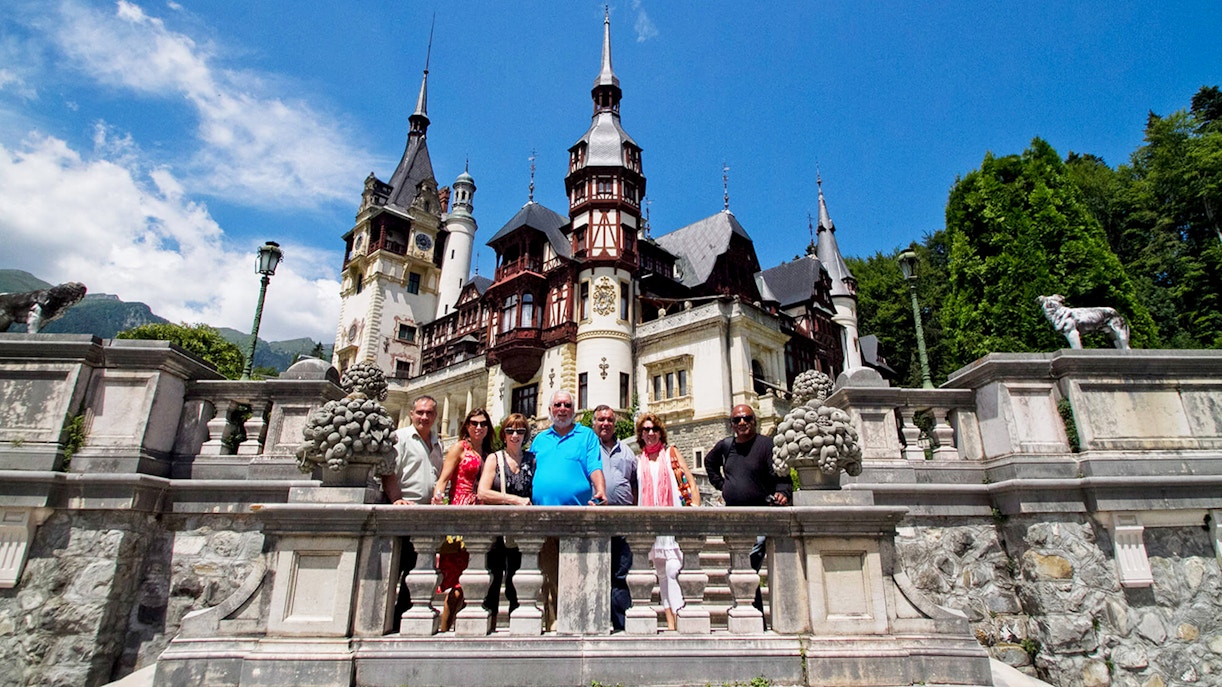 Guests standing in front of Peles Castle, Romania, with ornate architecture and spires in the background.