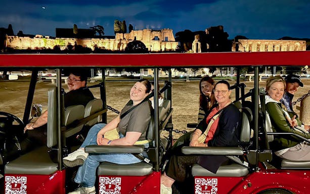 Golf cart tour group enjoying Rome by night with illuminated ancient ruins in the background.