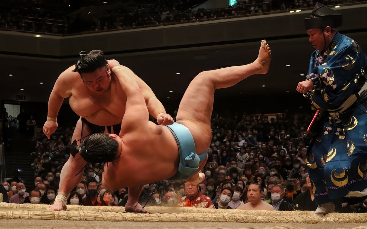 Sumo wrestler executing a move against opponent in a crowded arena.