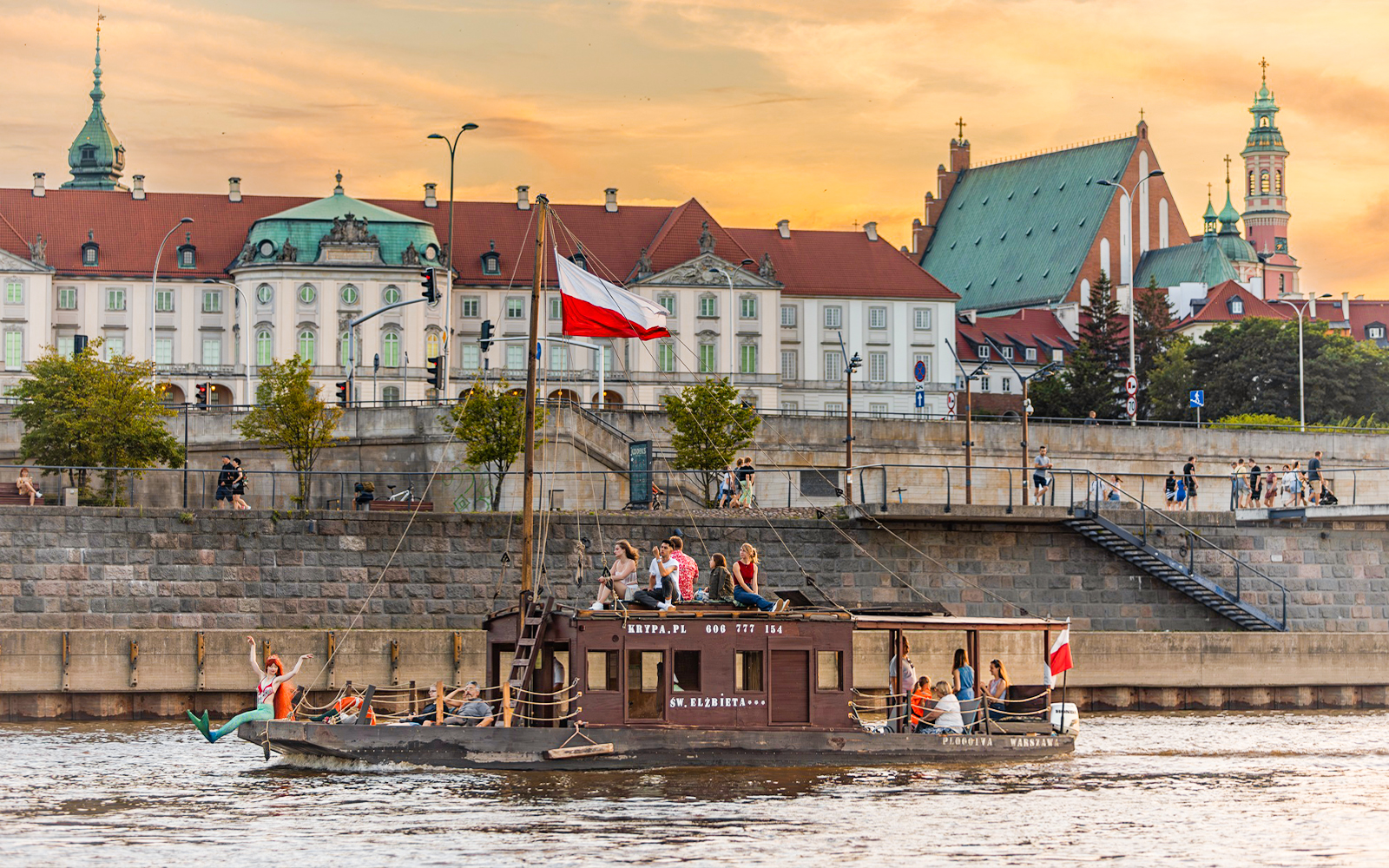 Tourists on a traditional Galar cruise on the Vistula River in Warsaw, Poland, with historic buildings in the background.