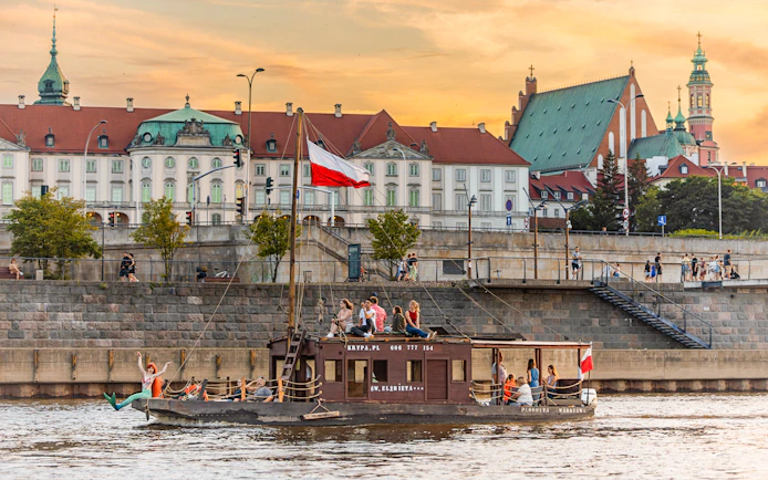 Tourists on a traditional Galar cruise on the Vistula River in Warsaw, Poland, with historic buildings in the background.