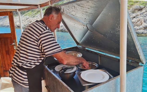 Man preparing food on a boat in Dubrovnik, with coastal scenery in the background.