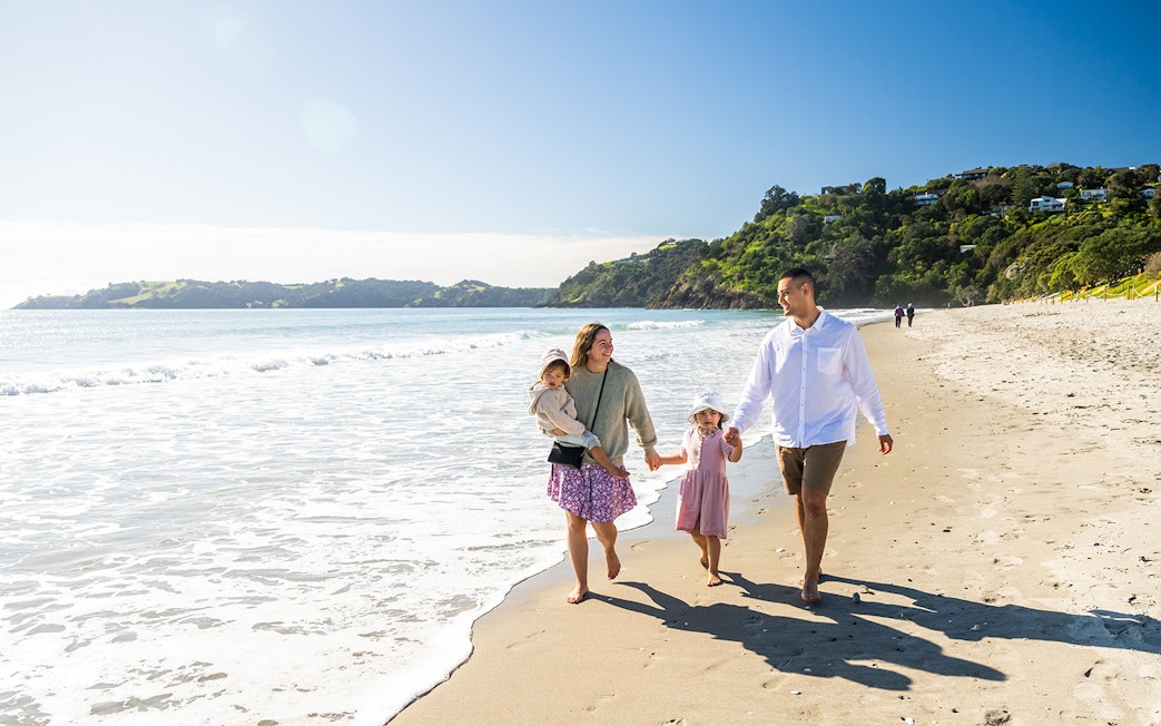 Family walking along Onetangi Beach, Waiheke Island, with lush hills in the background.