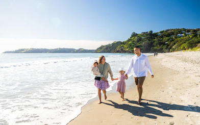 Family walking along Onetangi Beach, Waiheke Island, with lush hills in the background.