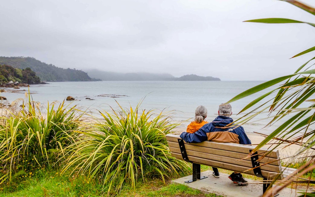Couple sitting on a bench overlooking the beach and bay on Stewart Island tour.
