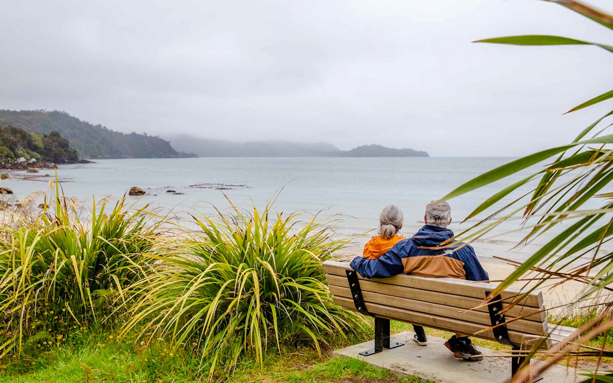 Couple sitting on a bench overlooking the beach and bay on Stewart Island tour.