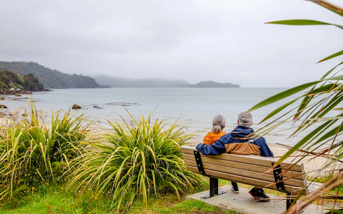 Couple sitting on a bench overlooking the beach and bay on Stewart Island tour.