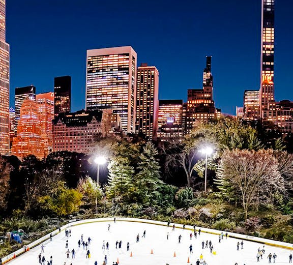 Skaters enjoying Wollman Ice Rink in Central Park with New York City skyline at night.