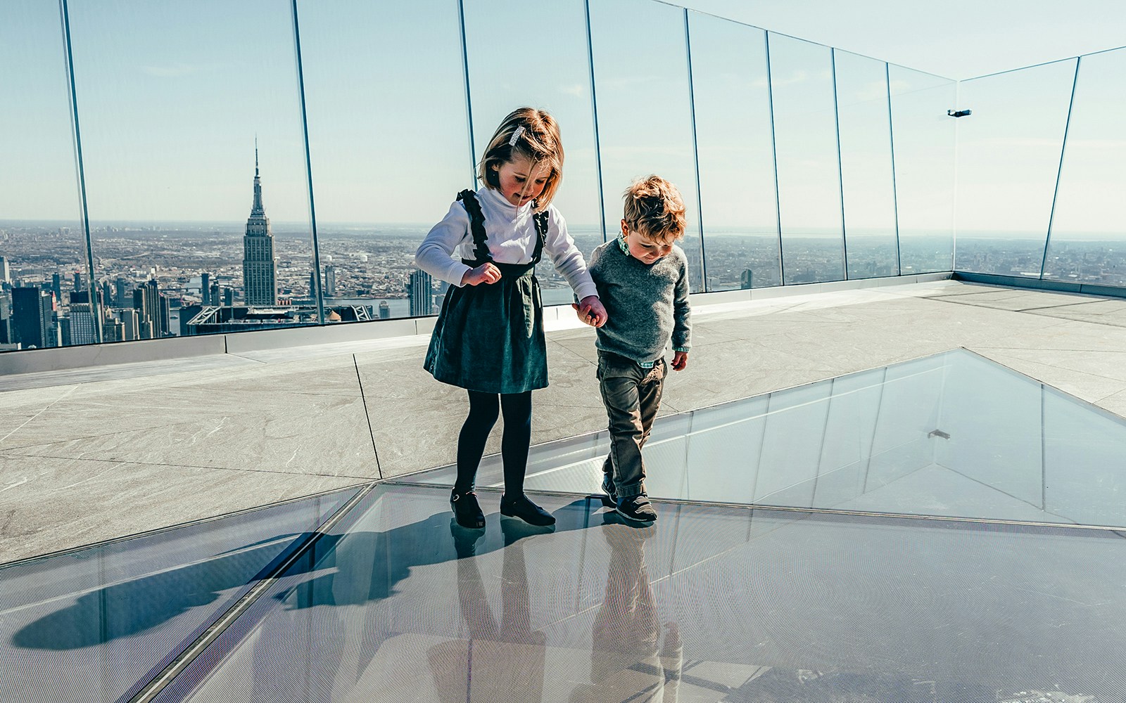 Children walking on glass floor at Edge observation deck, New York City skyline in background.