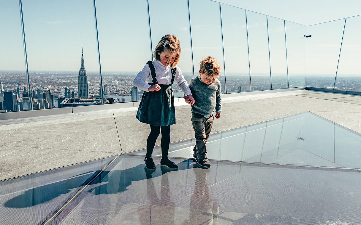 Children walking on glass floor at Edge observation deck, New York City skyline in background.