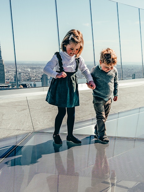 Children walking on glass floor at Edge observation deck, New York City skyline in background.