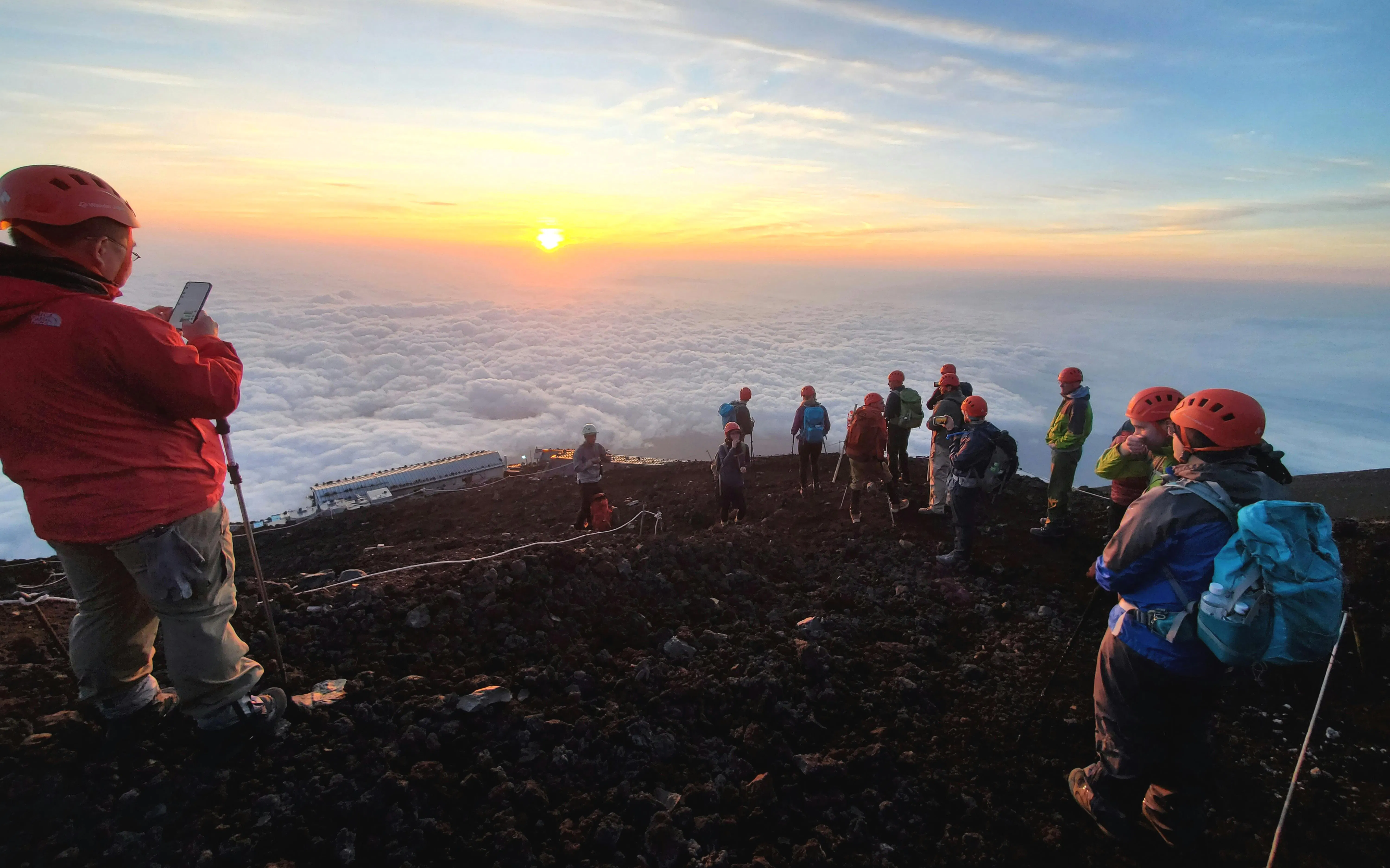 Climbers on Mt. Fuji summit at sunrise, Japan, with clouds below.