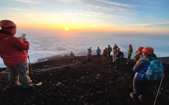 Climbers on Mt. Fuji summit at sunrise, Japan, with clouds below.