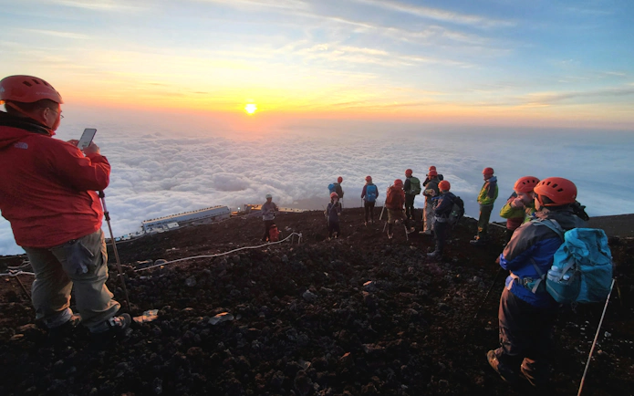 Climbers on Mt. Fuji summit at sunrise, Japan, with clouds below.