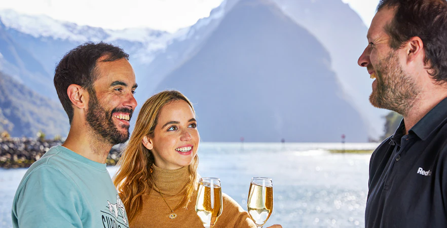 Couple enjoying drinks on Milford Sound overnight cruise with scenic mountain view.