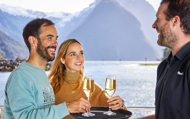Couple enjoying drinks on Milford Sound overnight cruise with scenic mountain view.