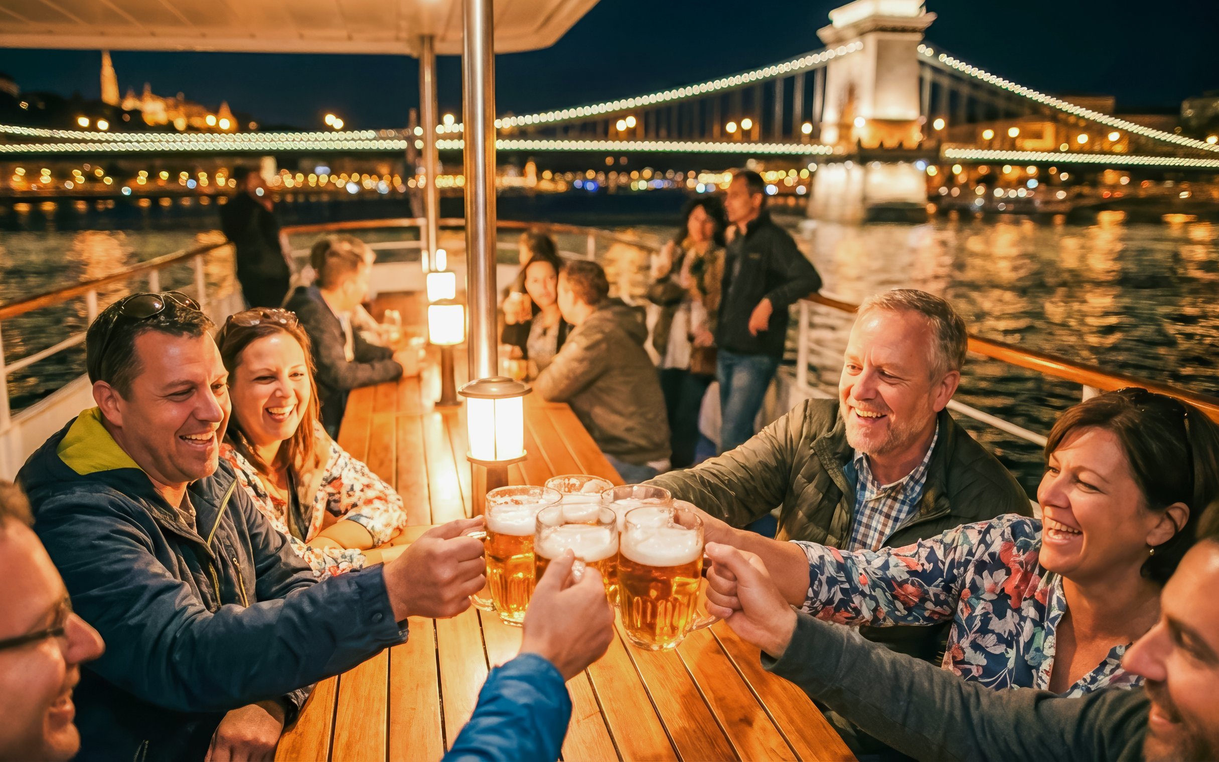 Guests toasting with beer on Budapest Danube River cruise with illuminated bridge in background.