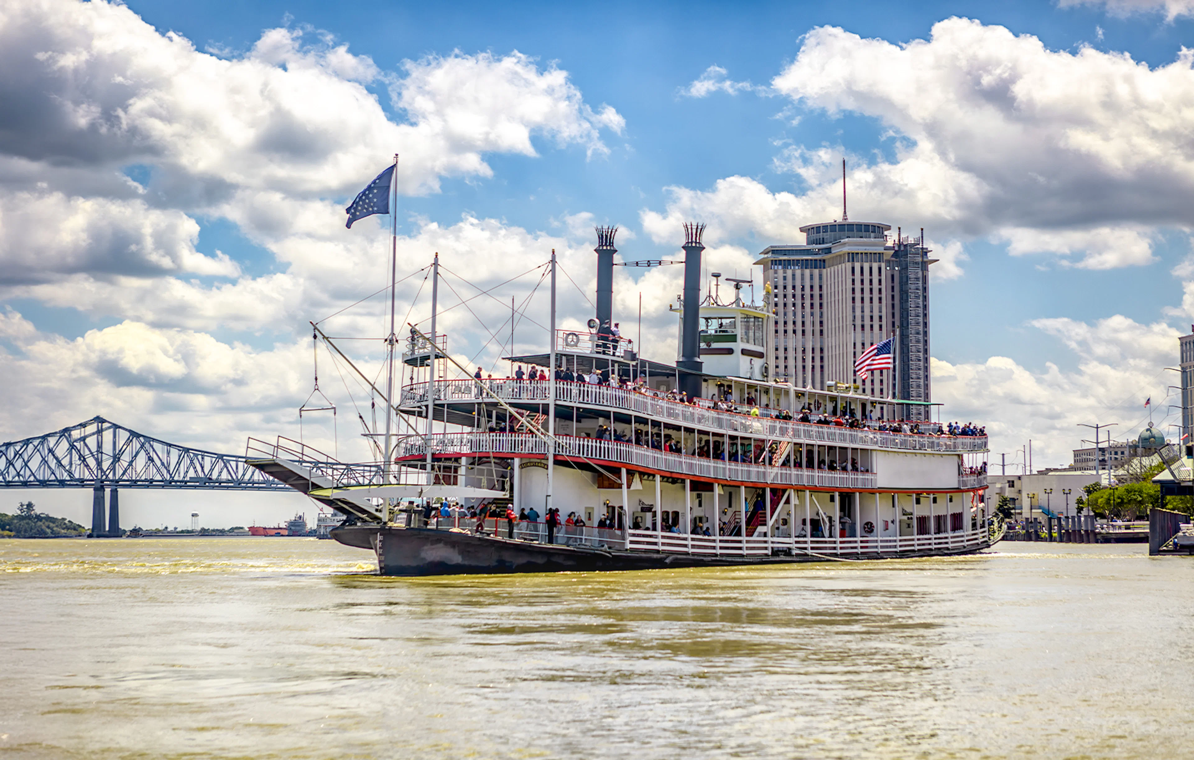 Steamboat Natchez cruising on the Mississippi River in New Orleans, Louisiana, with city skyline and bridge in view.