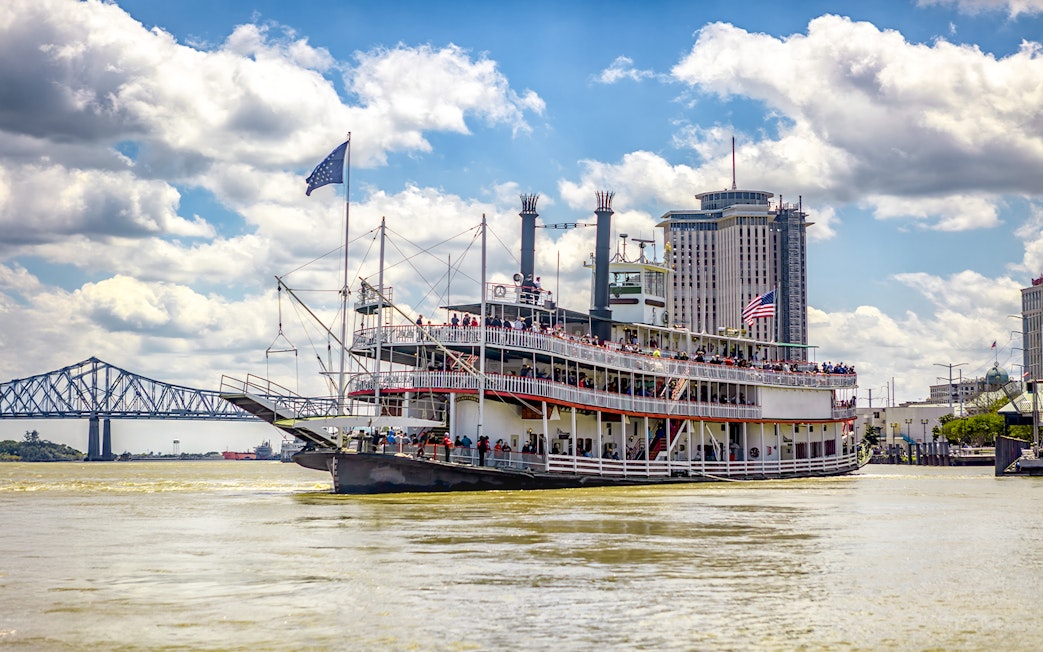 Steamboat Natchez cruising on the Mississippi River in New Orleans, Louisiana, with city skyline and bridge in view.