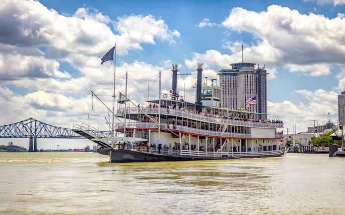 Steamboat Natchez cruising on the Mississippi River in New Orleans, Louisiana, with city skyline and bridge in view.