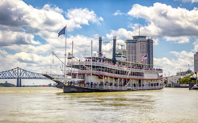 Steamboat Natchez cruising on the Mississippi River in New Orleans, Louisiana, with city skyline and bridge in view.