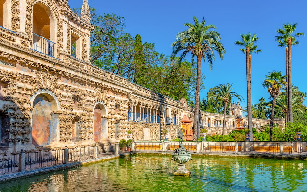 Mercury fountain in the gardens of the Real Alcazar de Sevilla, Spain, with palm trees and historic architecture.