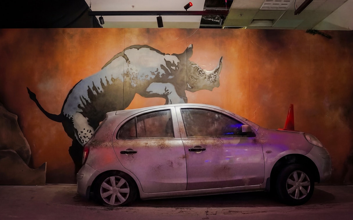 Rhino mural on wall behind parked car, Banksy Limitless exhibit.