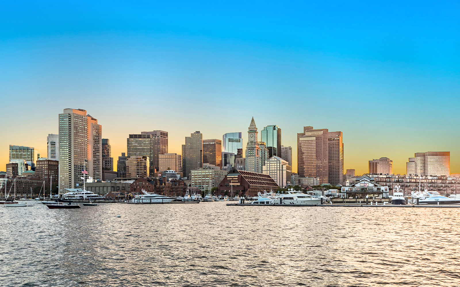 Boston skyline at sunset with waterfront and boats in the foreground.