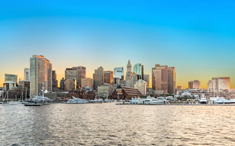 Boston skyline at sunset with waterfront and boats in the foreground.