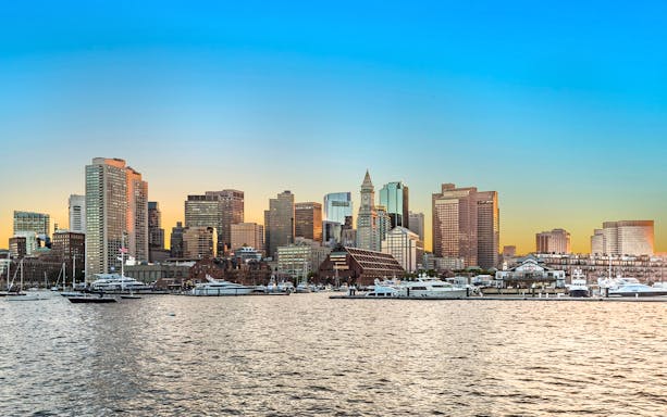 Boston skyline at sunset with waterfront and boats in the foreground.