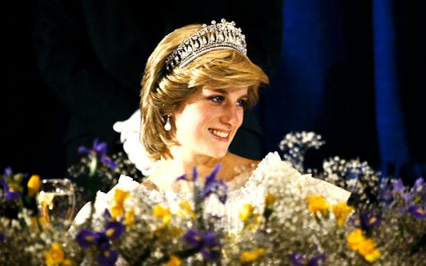 Princess Diana smiling at a formal event, wearing a tiara, with flowers in the foreground.