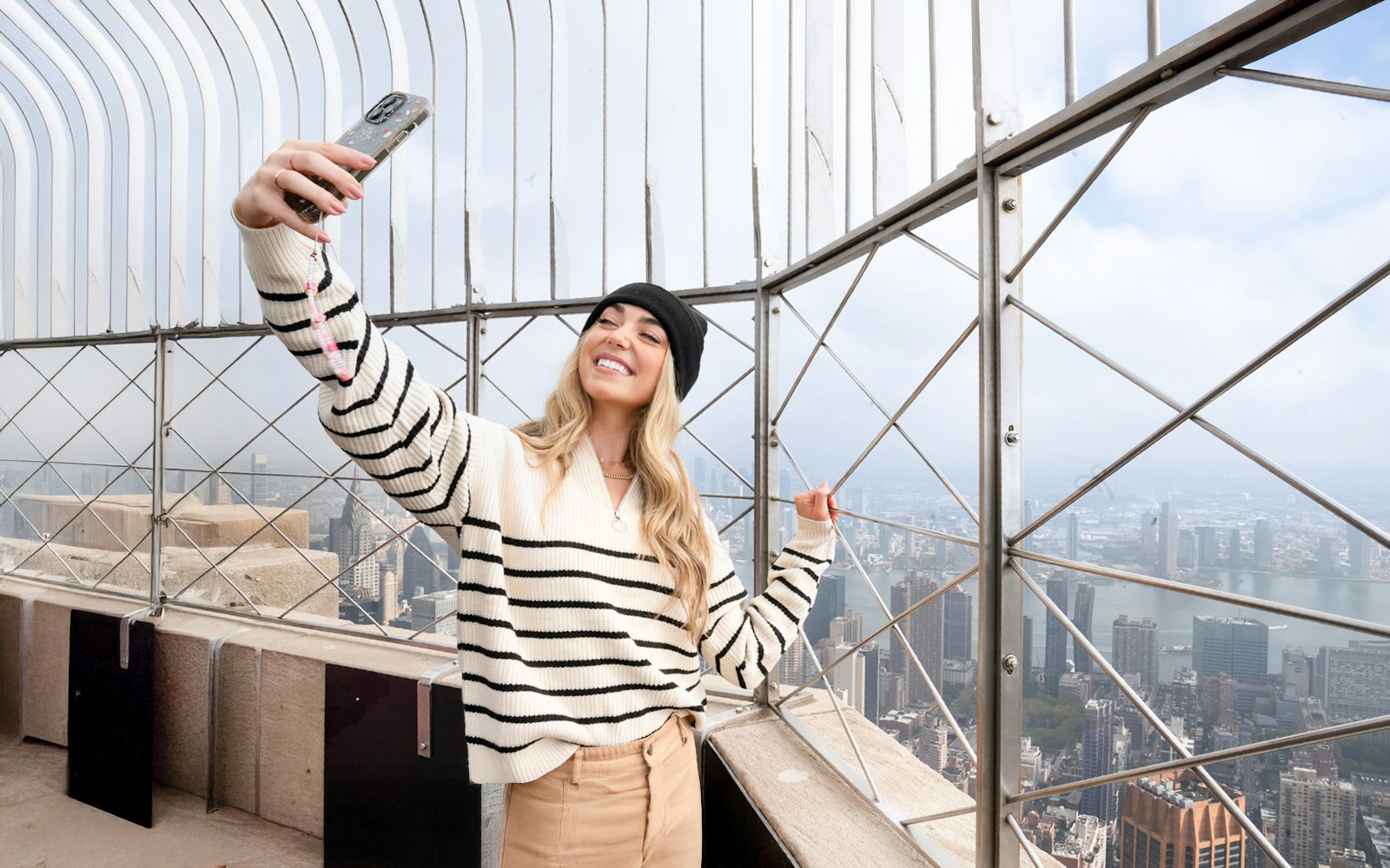 Woman taking a selfie on the Empire State Building observation deck, NYC skyline in background.