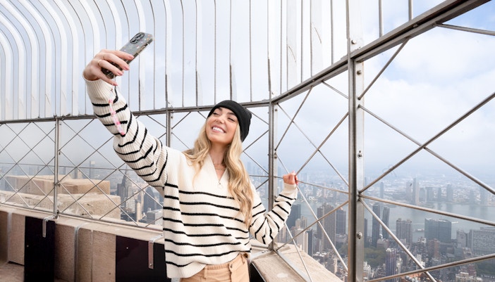 Woman taking a selfie on the Empire State Building observation deck, NYC skyline in background.