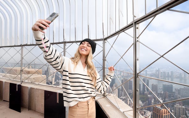 Woman taking a selfie on the Empire State Building observation deck, NYC skyline in background.