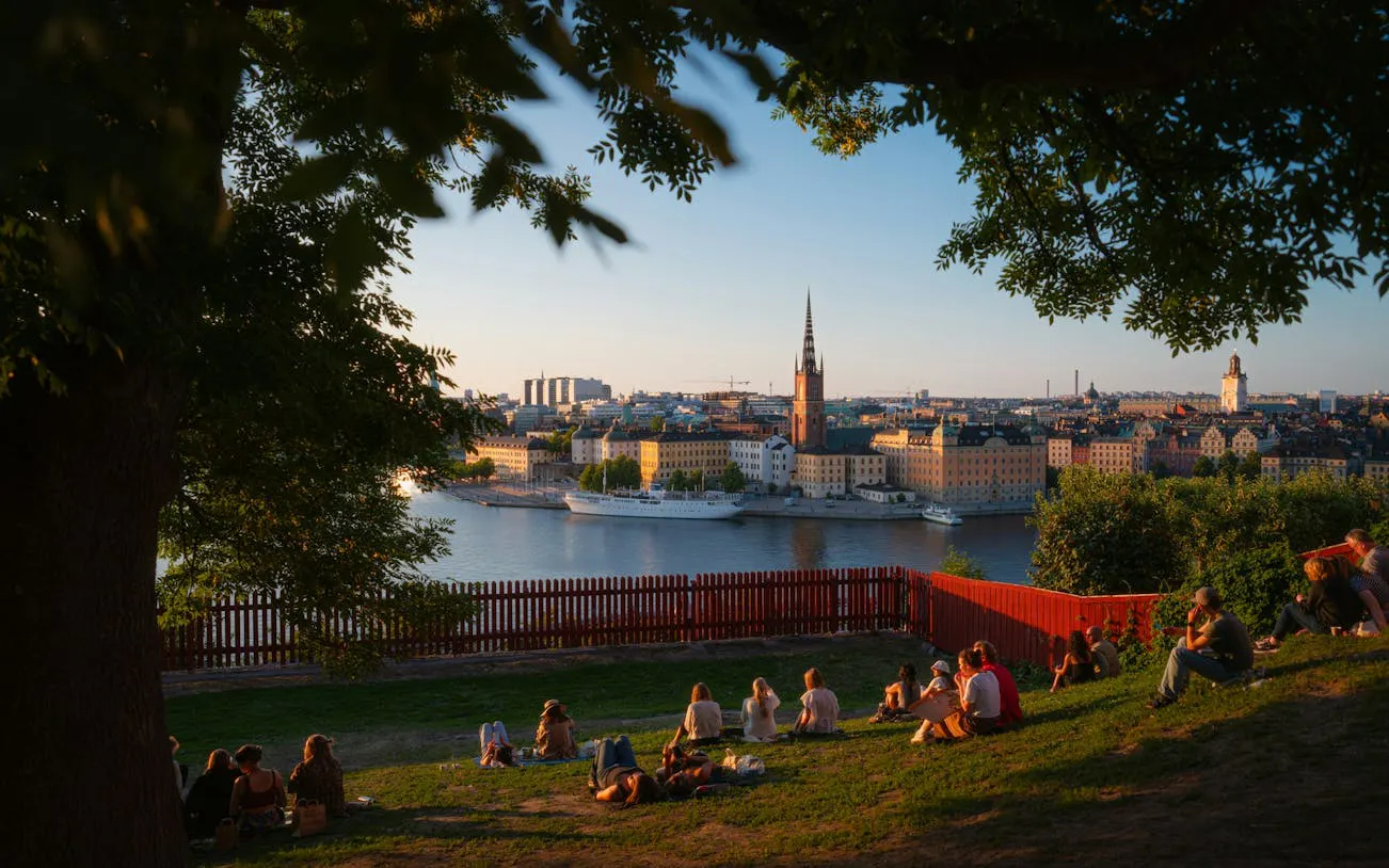 People relaxing on a grassy hill overlooking Stockholm's waterfront and cityscape at sunset.