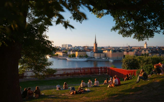 People relaxing on a grassy hill overlooking Stockholm's waterfront and cityscape at sunset.