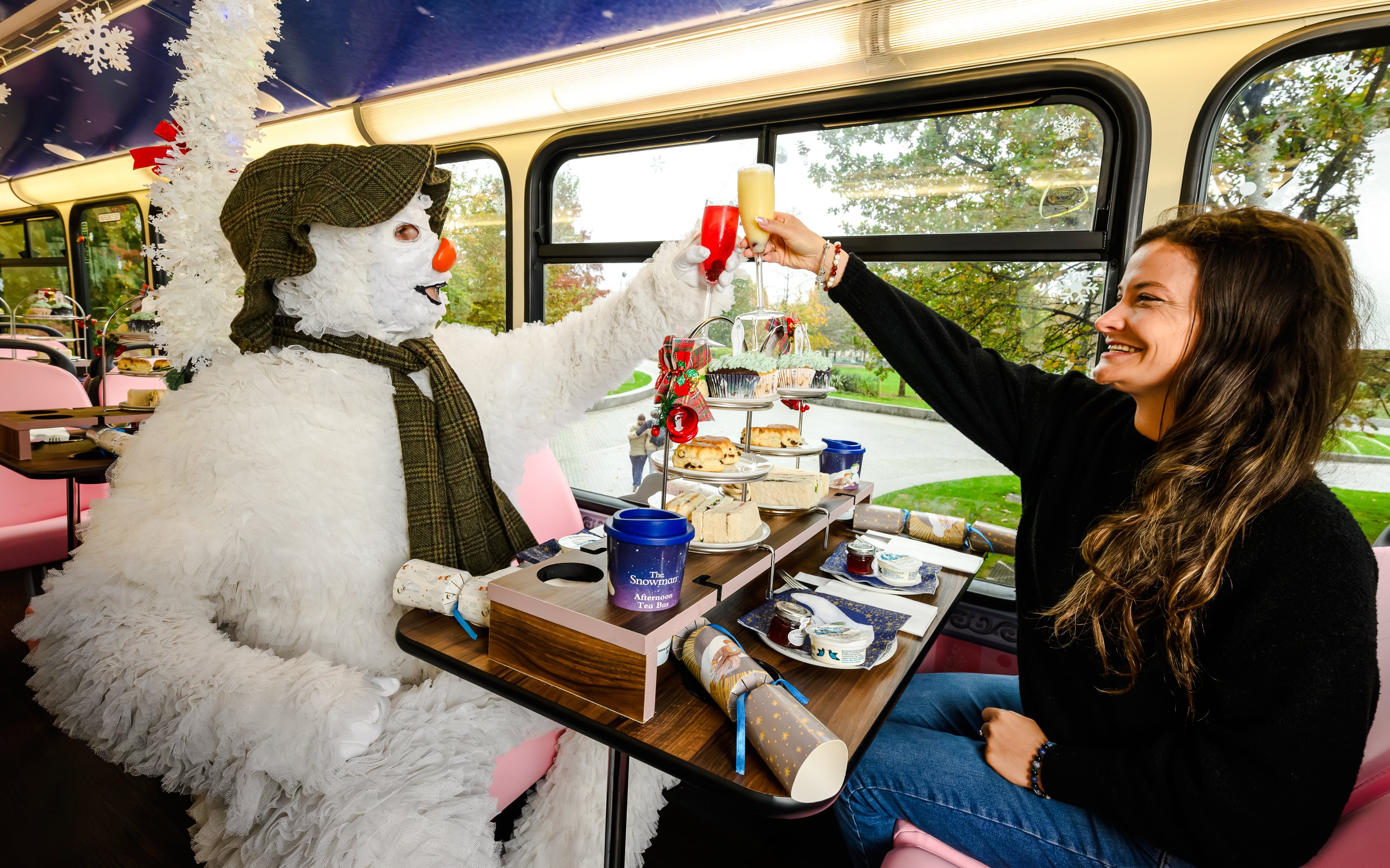 Snowman character and woman toasting on The Snowman™ Afternoon Tea Bus Tour with festive treats.