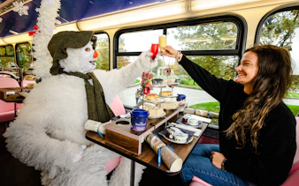 Snowman character and woman toasting on The Snowman™ Afternoon Tea Bus Tour with festive treats.