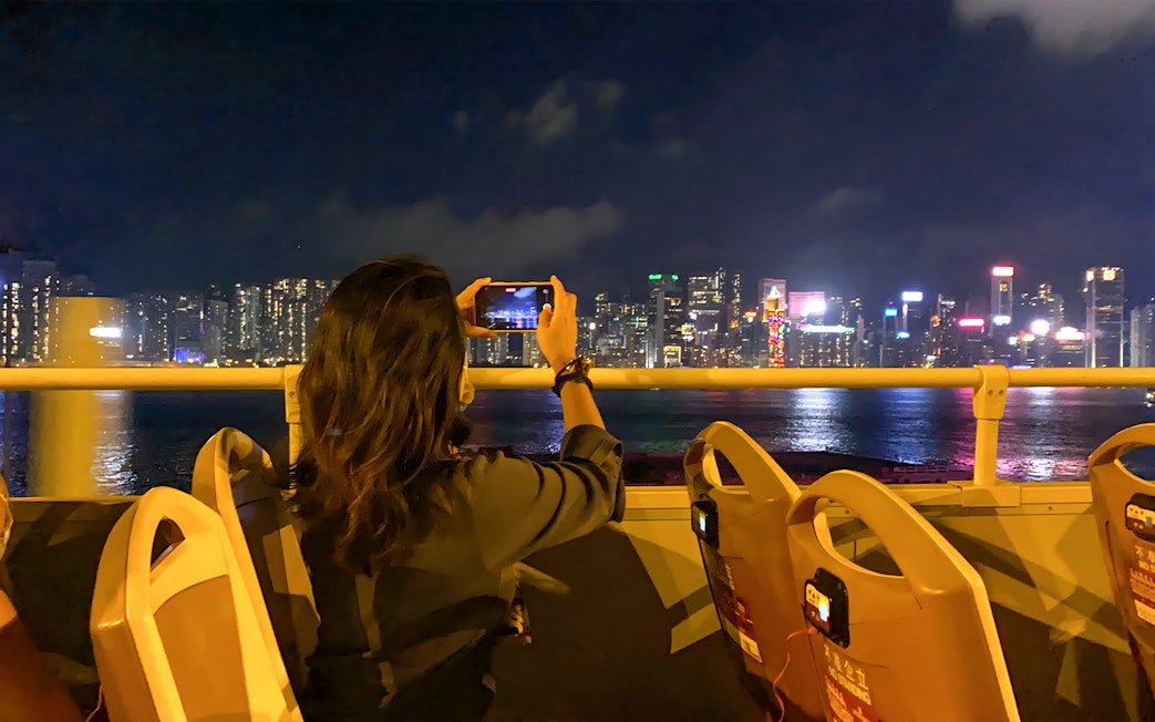 Person taking photo of Hong Kong skyline from open-top bus at night.