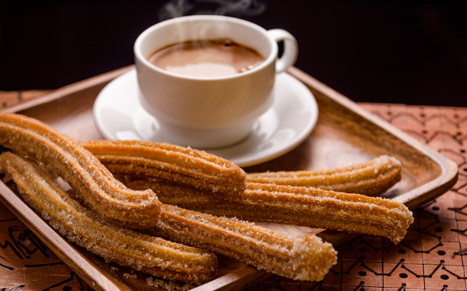 Churros with sugar on a wooden tray beside a steaming cup of chocolate.
