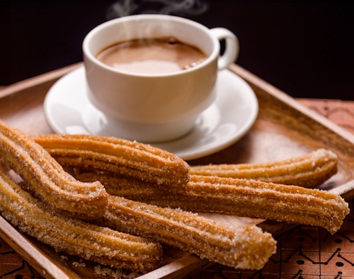 Churros with sugar on a wooden tray beside a steaming cup of chocolate.