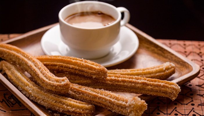 Churros with sugar on a wooden tray beside a steaming cup of chocolate.