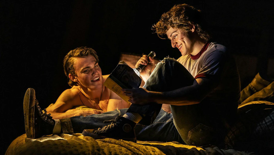 Two actors reading a book with a flashlight during The Outsiders stage performance on Broadway.