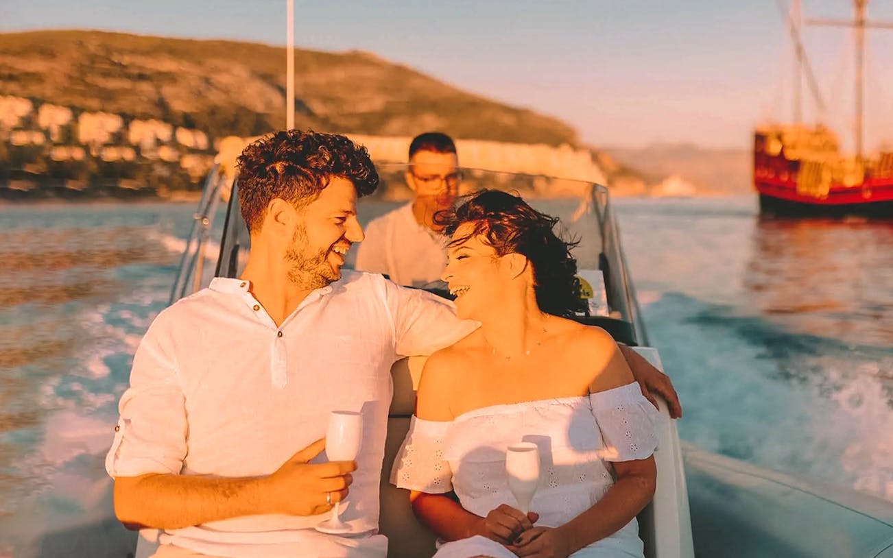 Guests on a private boat cruise with drinks, viewing the sunset over Dubrovnik's coastline.