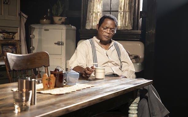 Man sitting at a wooden table with a mug, scene from "The Piano Lesson" play.