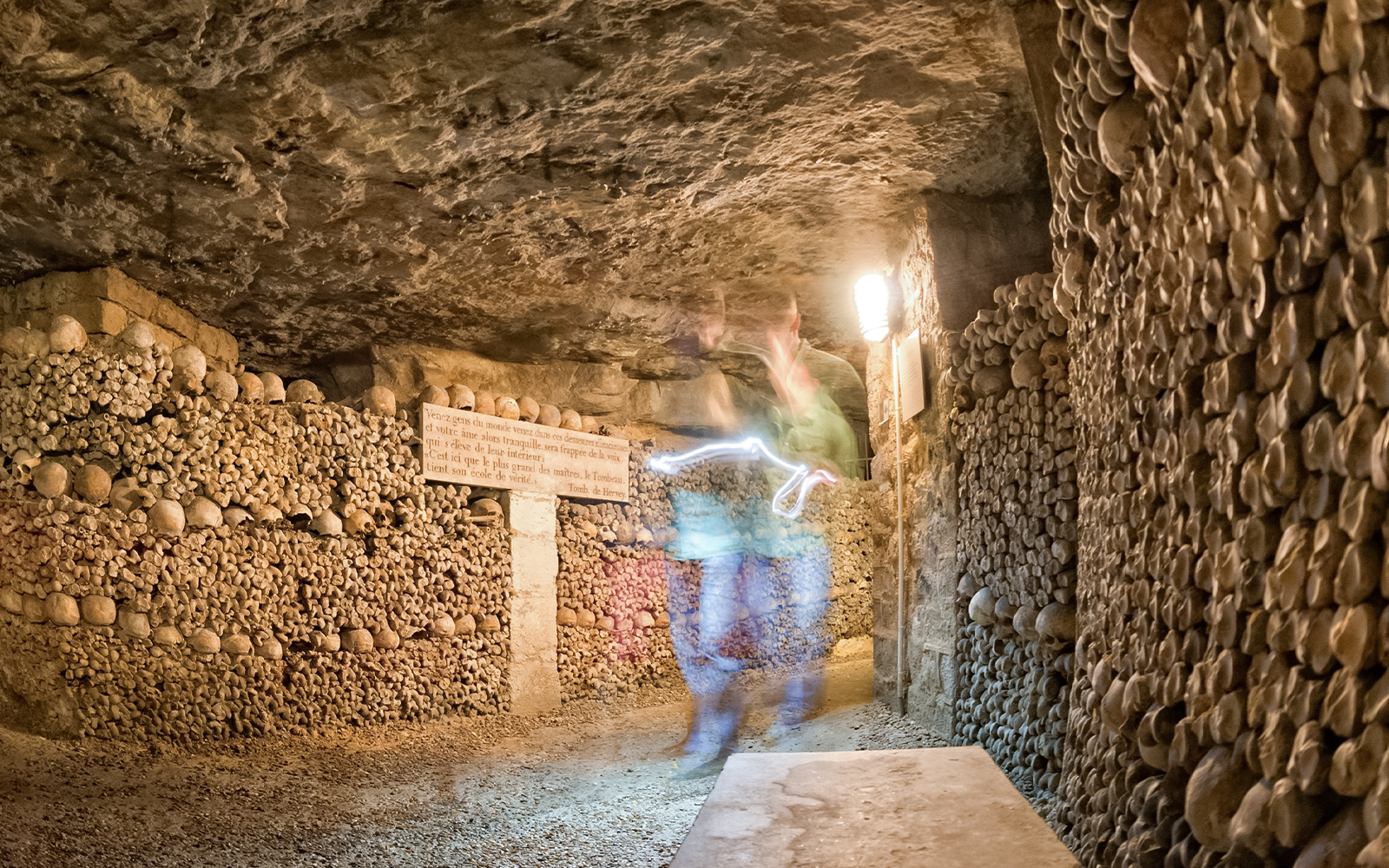 Ghostly figure in Paris Catacombs corridor with skull-lined walls.