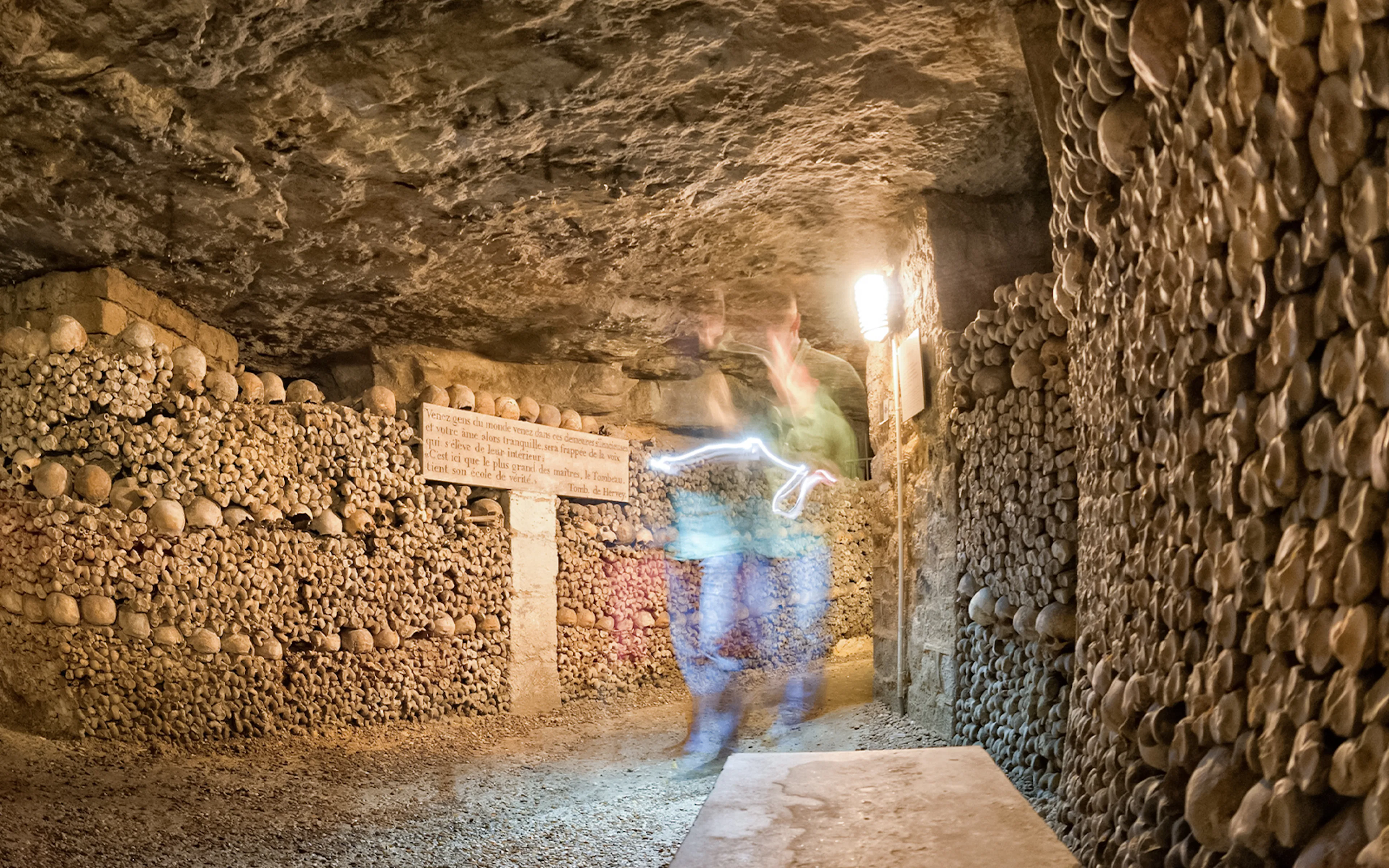 Ghostly figure in Paris Catacombs corridor with skull-lined walls.