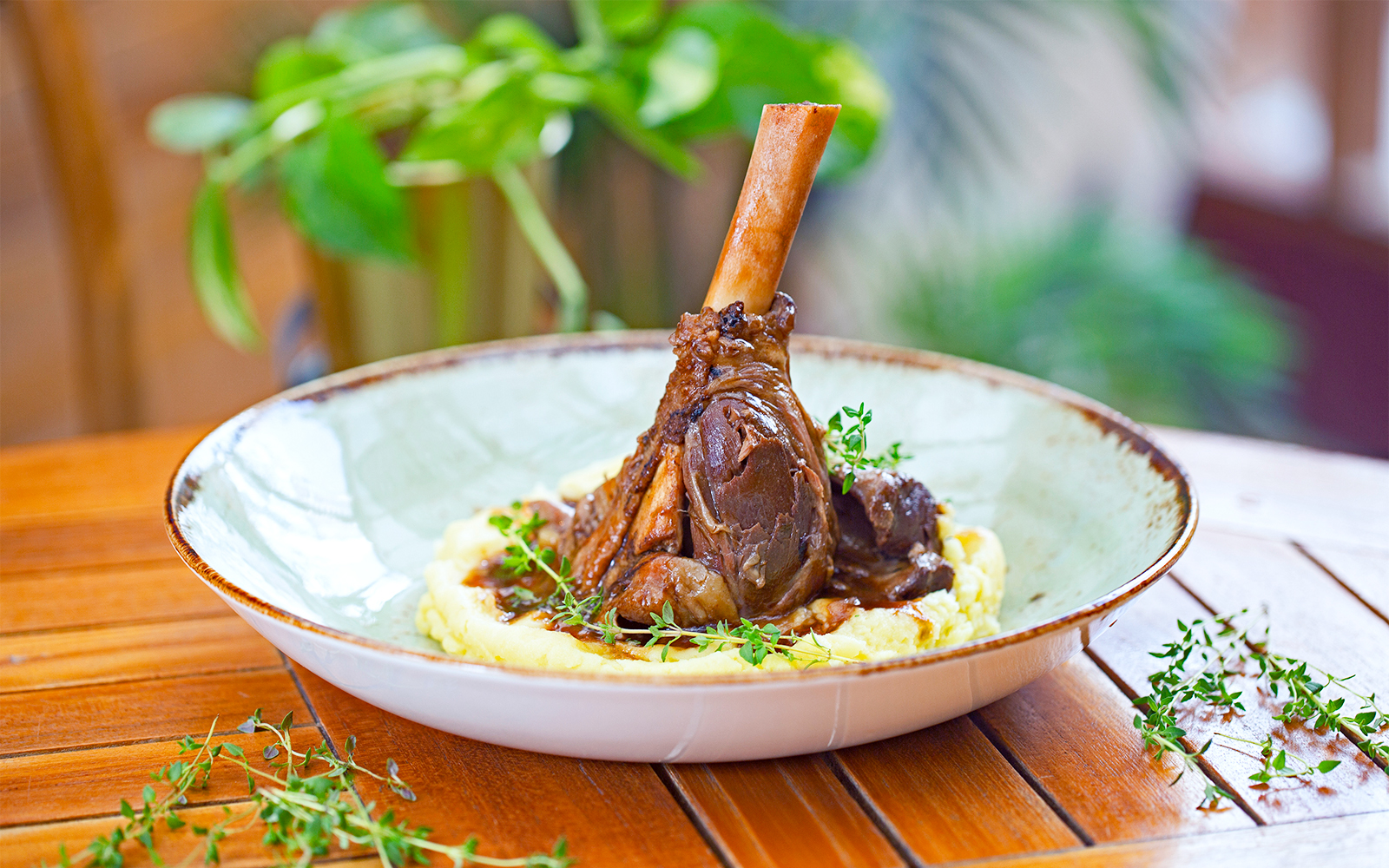 Braised lamb shank on mashed potatoes with herbs, served on a wooden table.