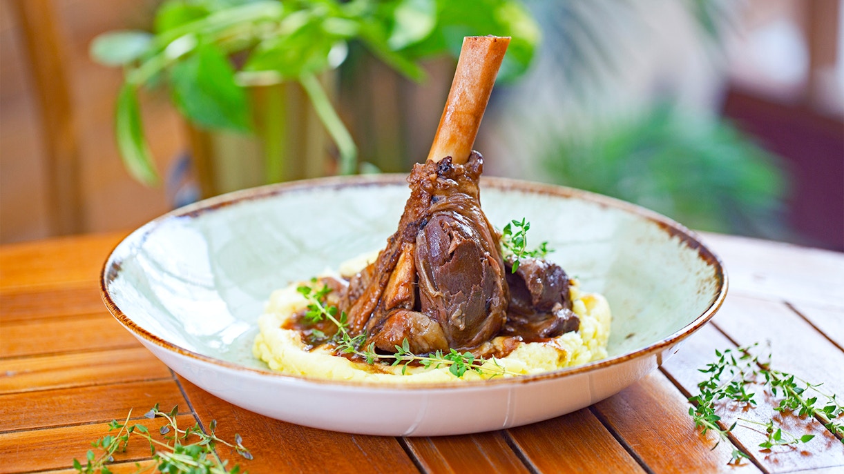 Braised lamb shank on mashed potatoes with herbs, served on a wooden table.